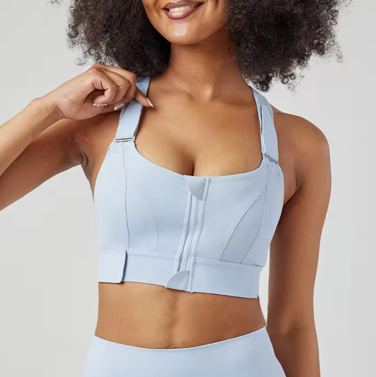 Femme souriante portant une brassière de sport bleu clair avec fermeture éclair, design moderne et confortable, idéale pour fitness et yoga.